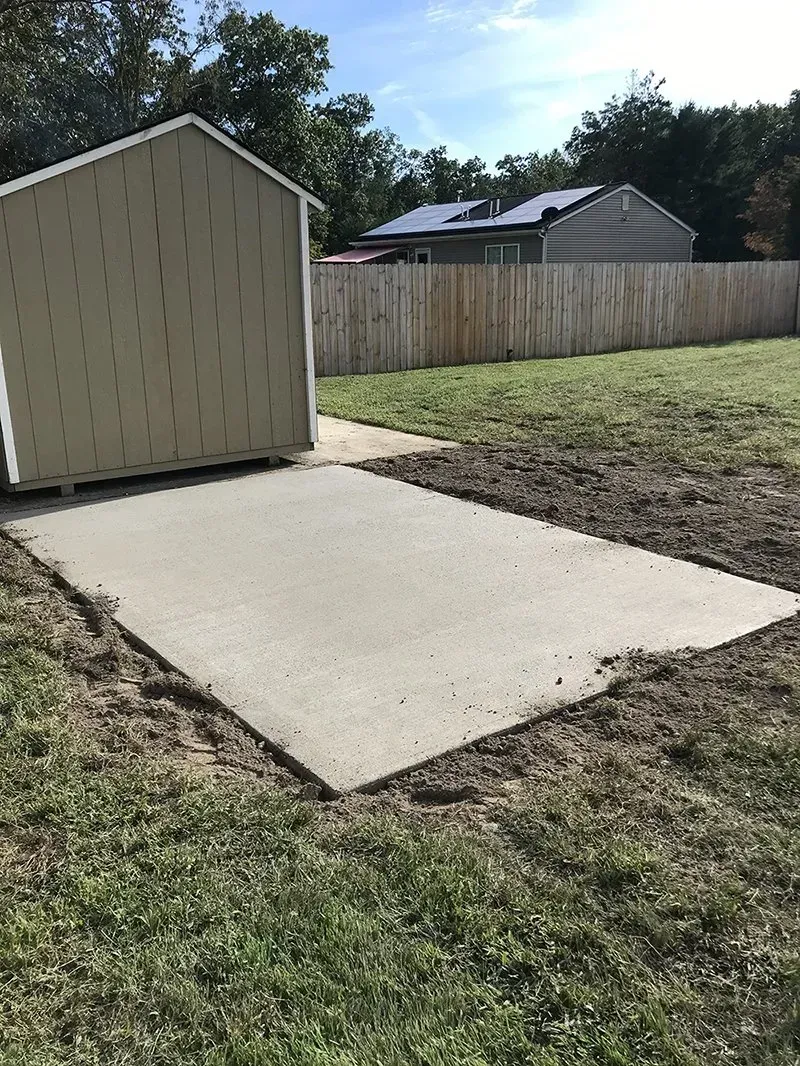 Concrete slab beside a beige shed in a grassy backyard, with a wooden fence and house in the background