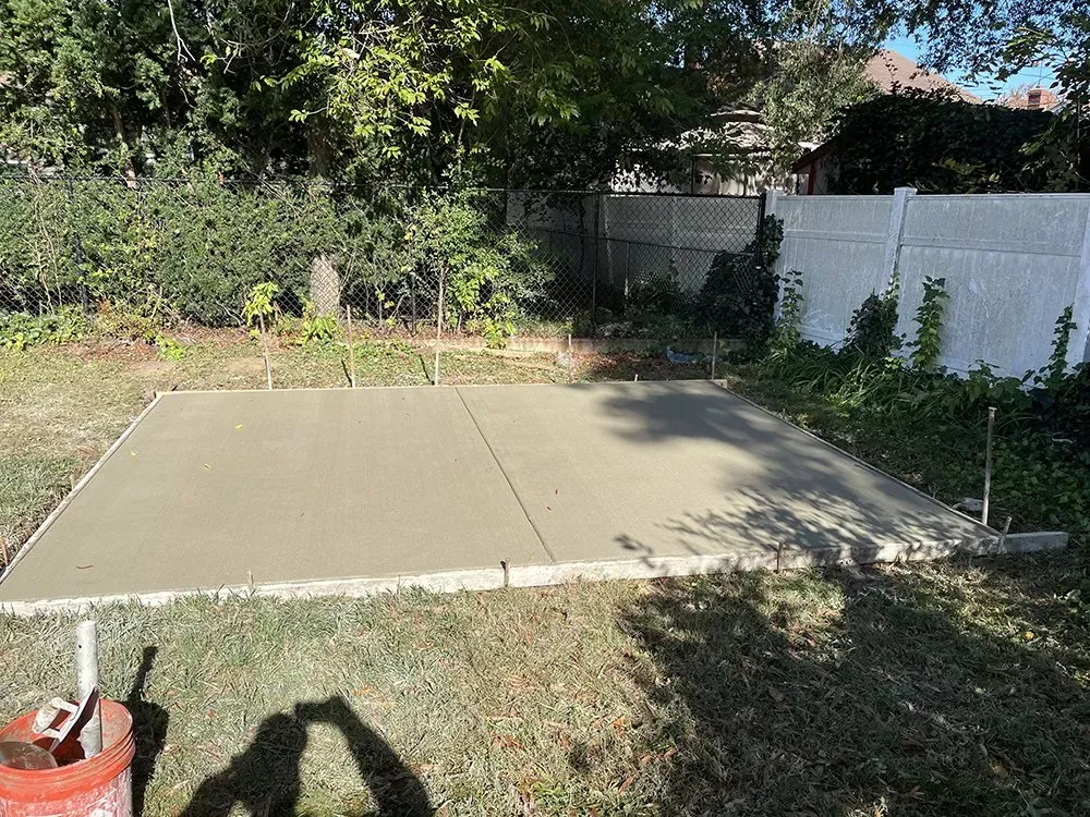 Concrete slab patio in a backyard, bordered by grass, trees, and a white fence.