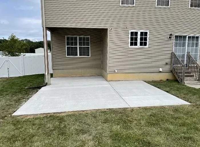 Concrete driveway and covered patio beside a beige house with a white fence and grassy yard.