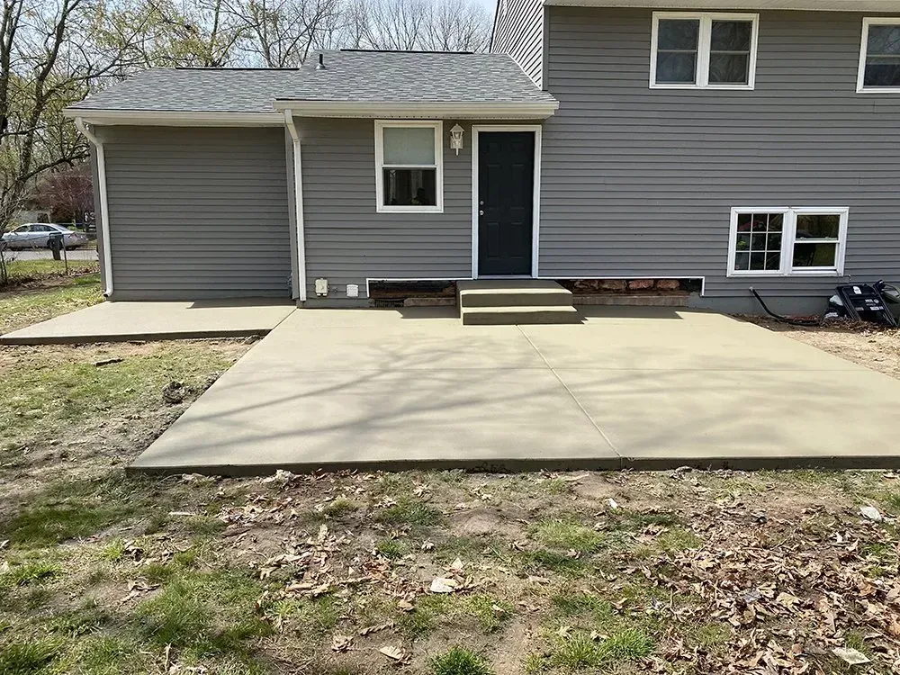 Concrete patio beside a gray house with a small porch and steps leading to a blue door