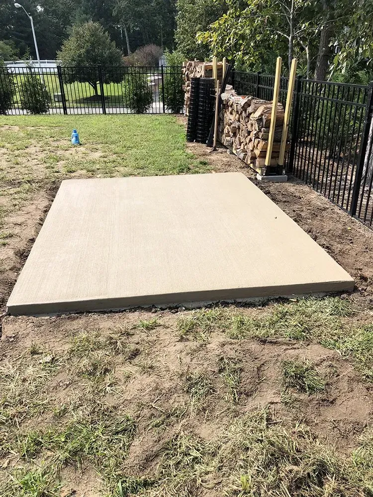 Concrete patio slab in a backyard beside a black fence and stacked firewood.