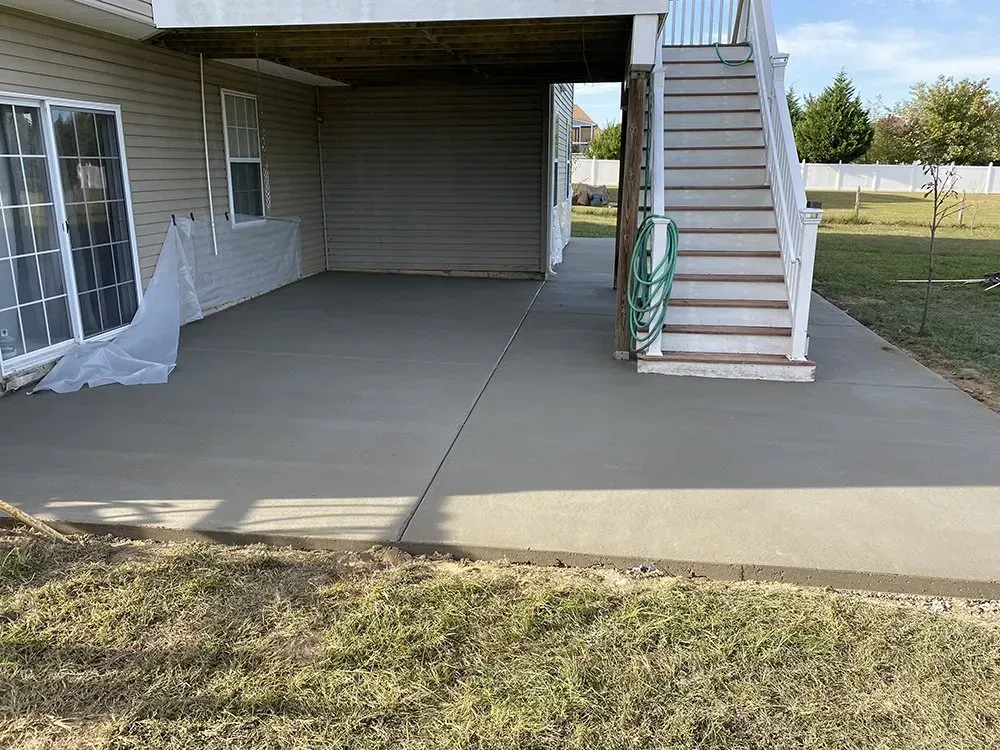 Concrete patio under a deck with stairs, sliding glass door, and grassy yard beyond.