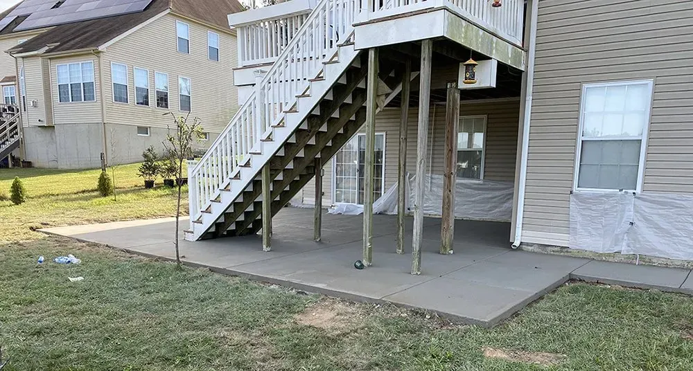 Elevated house deck with stairs and support posts over a concrete patio and grassy yard