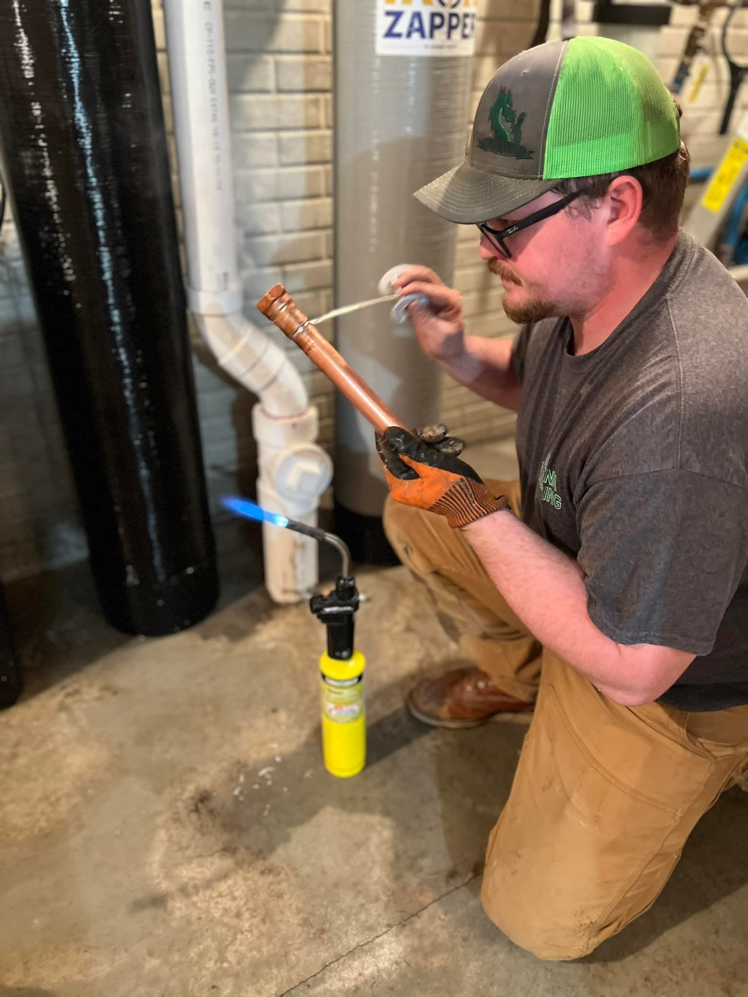 Plumber using a torch to solder copper pipe, wearing a cap and gloves, in a utility room.
