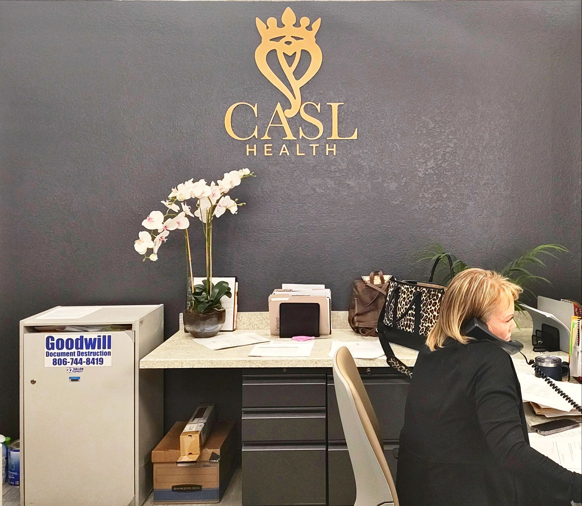 Reception desk with a woman on the phone under a gold CASL Health logo, and an artificial flower display.