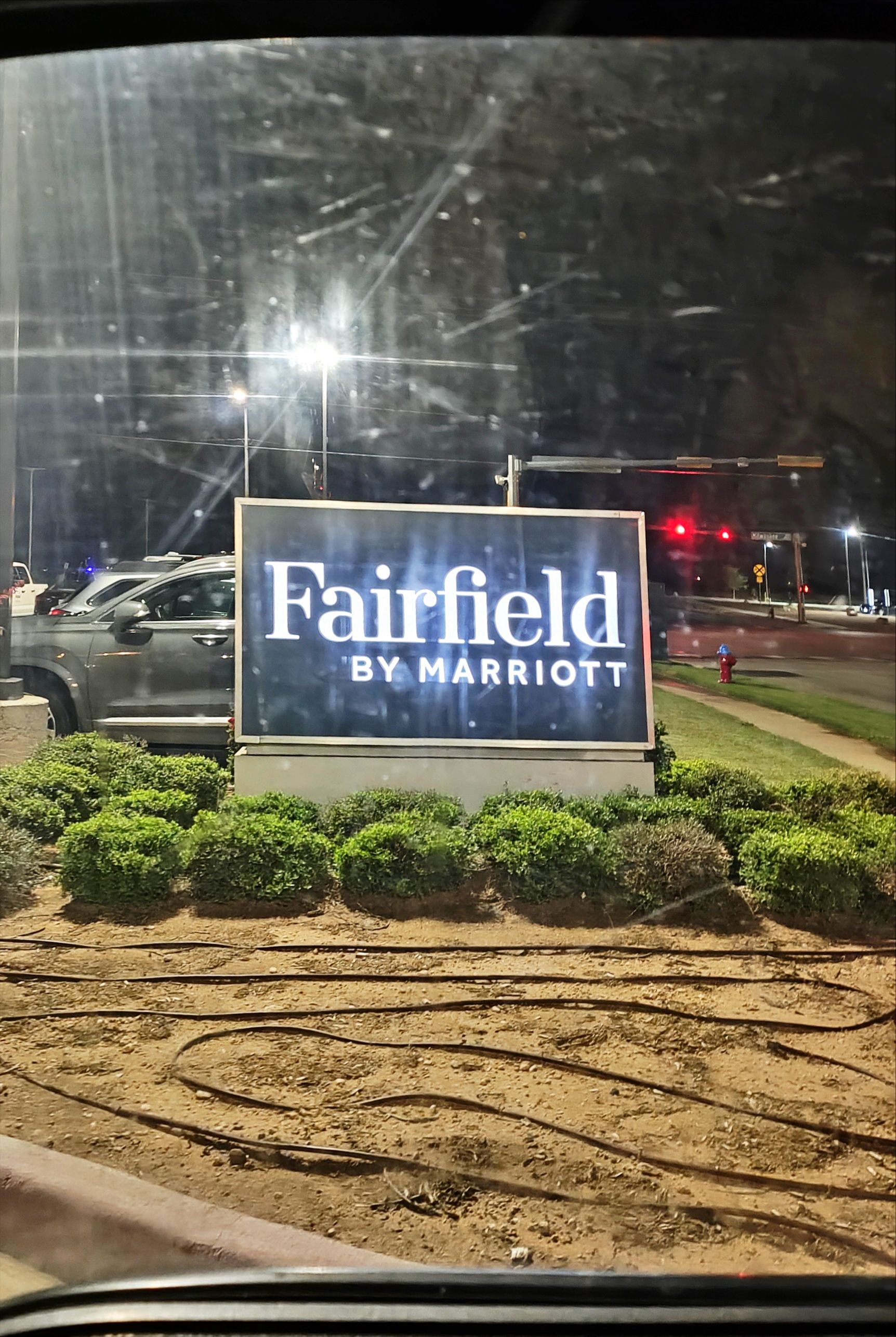 Fairfield by Marriott sign illuminated at night, bushes in front, cars and street visible in background.