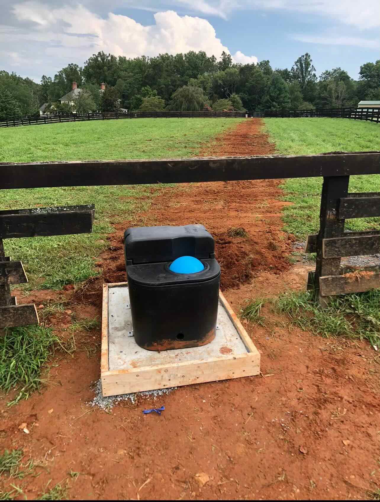 A black water tank is sitting in the dirt next to a wooden fence in a field.