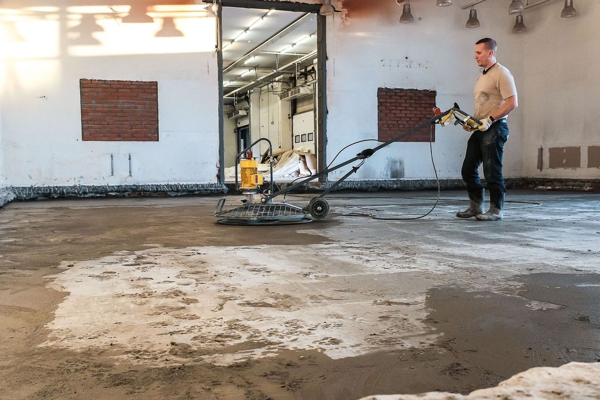 Man operating a power trowel, smoothing concrete floor in an industrial building with open doorway and brick-covered windows.