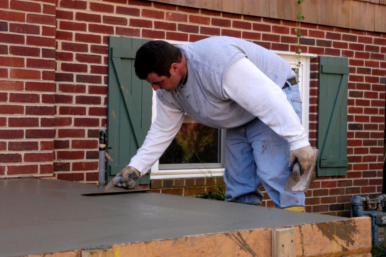 Man smoothing wet concrete with a trowel on a rectangular surface near a brick building.