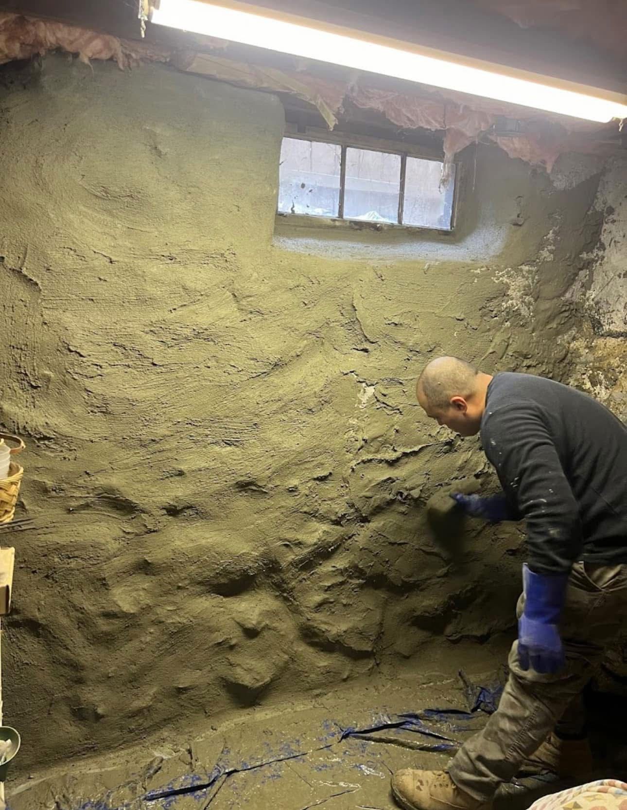 Person applying plaster to a stone wall in a basement, near a small window.