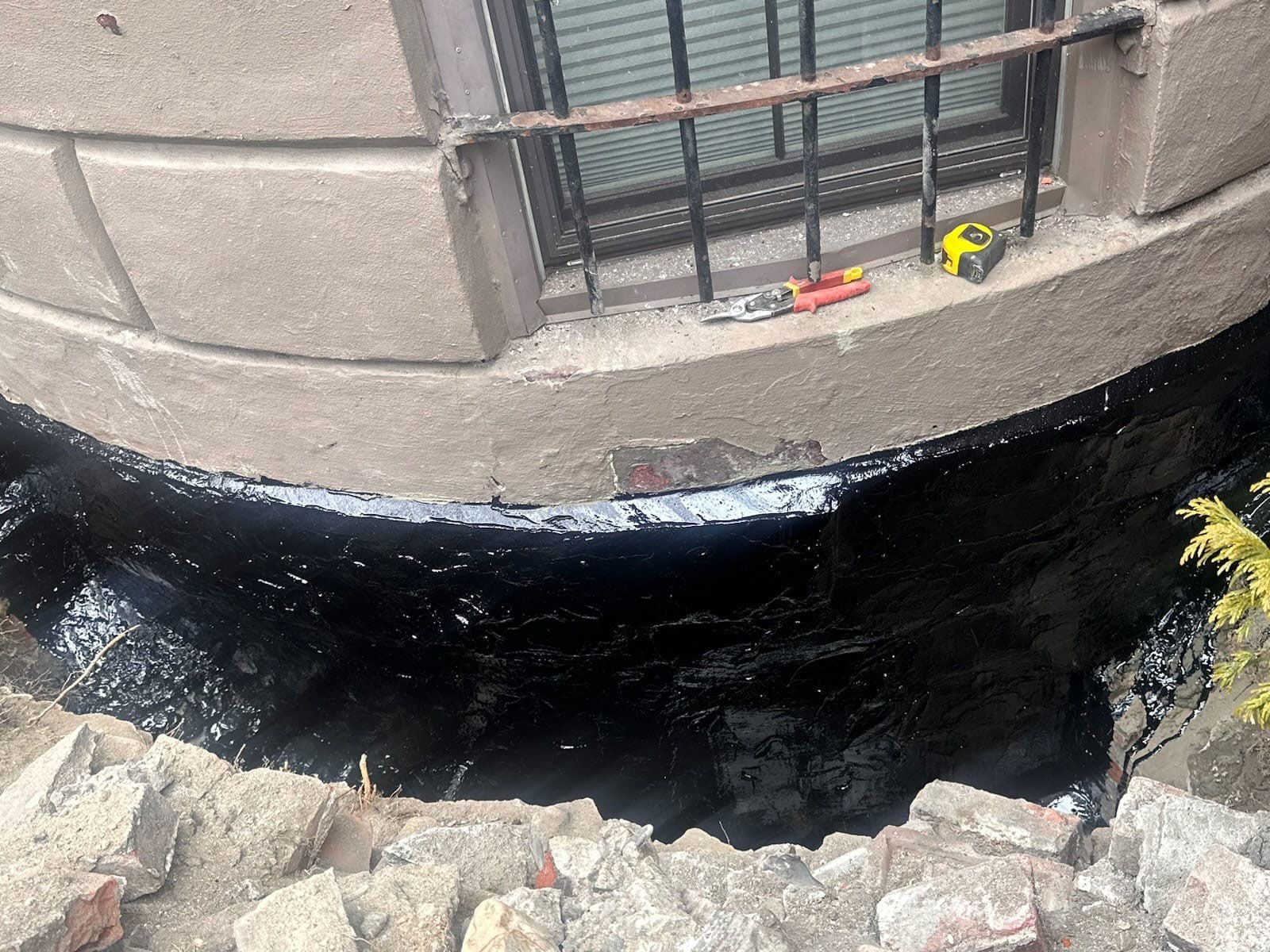 Black sealant applied to a concrete window well. Bricks and tools visible.