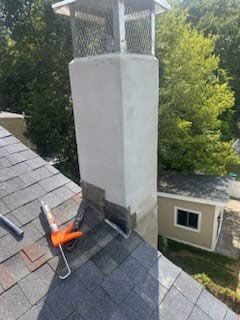 Chimney on a roof; gray and white structure with a cap, surrounded by shingles and trees.