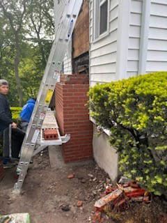 Workers repair brick chimney on house, using a ladder. Bricks and debris scattered on the ground.