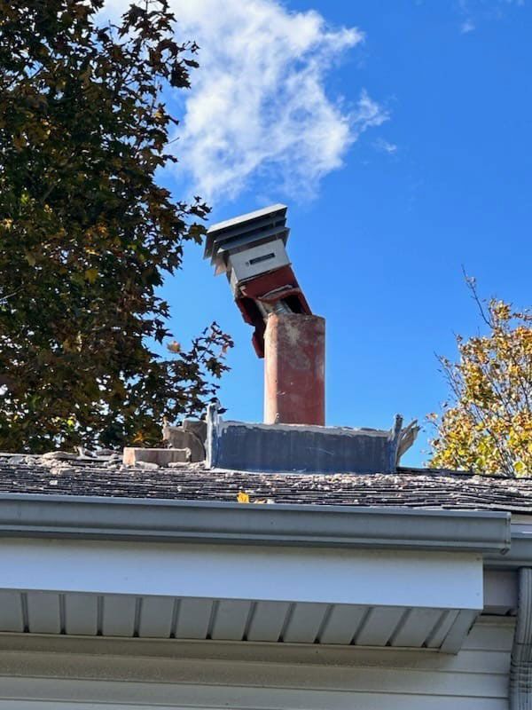 Damaged brick chimney on a roof, leaning at an angle. Blue sky and tree visible.