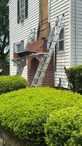 Workers rebuilding a brick chimney, using ladders and scaffolding on a two-story white house.