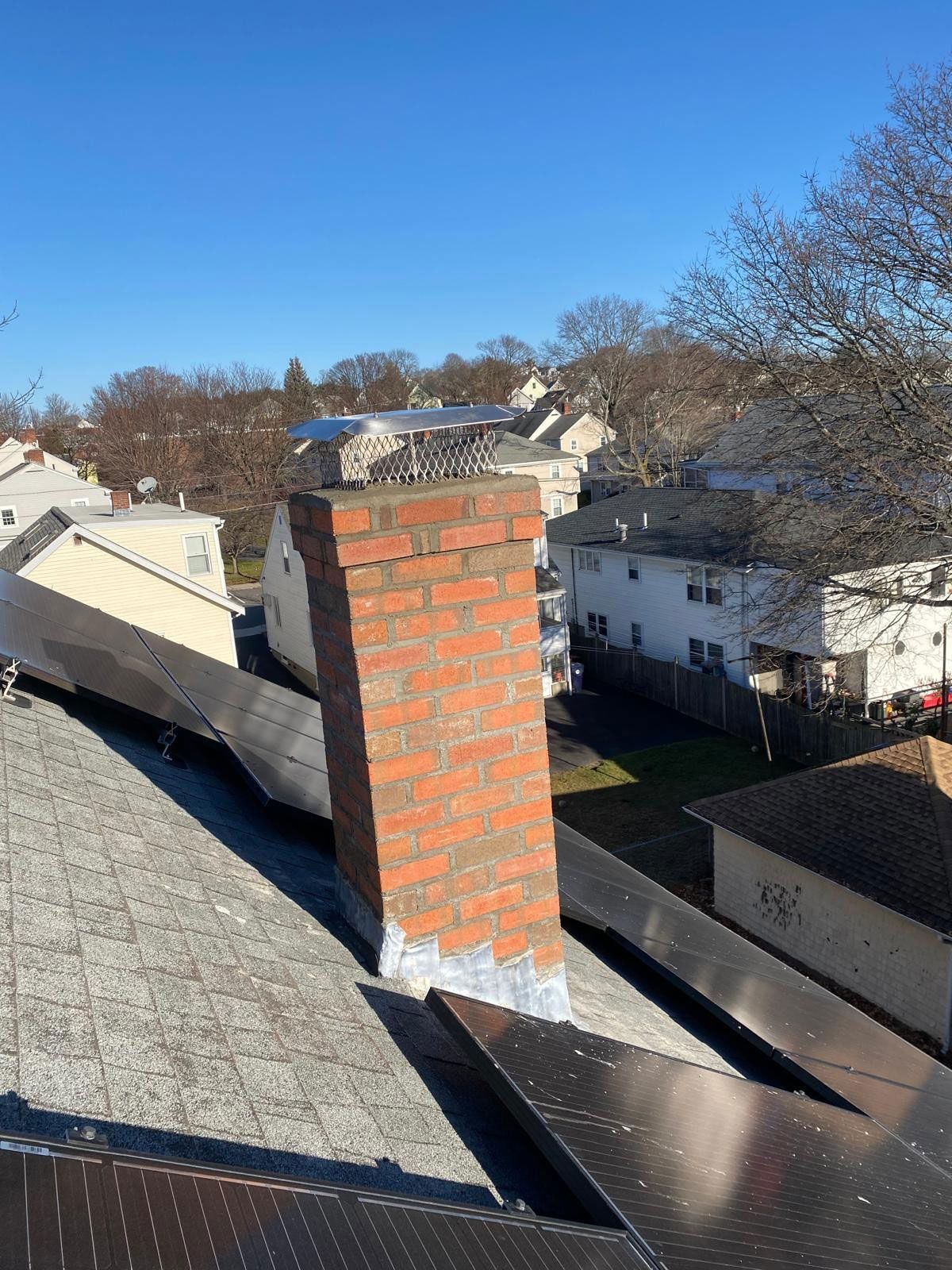 Leaning brick chimney on a rooftop with solar panels, houses in the background, clear blue sky.