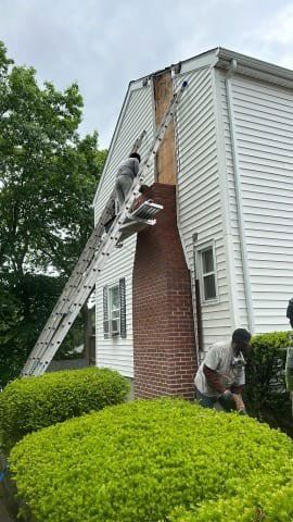 Workers on a ladder repairing a brick chimney on a two-story white house. One worker high up, another at ground level.