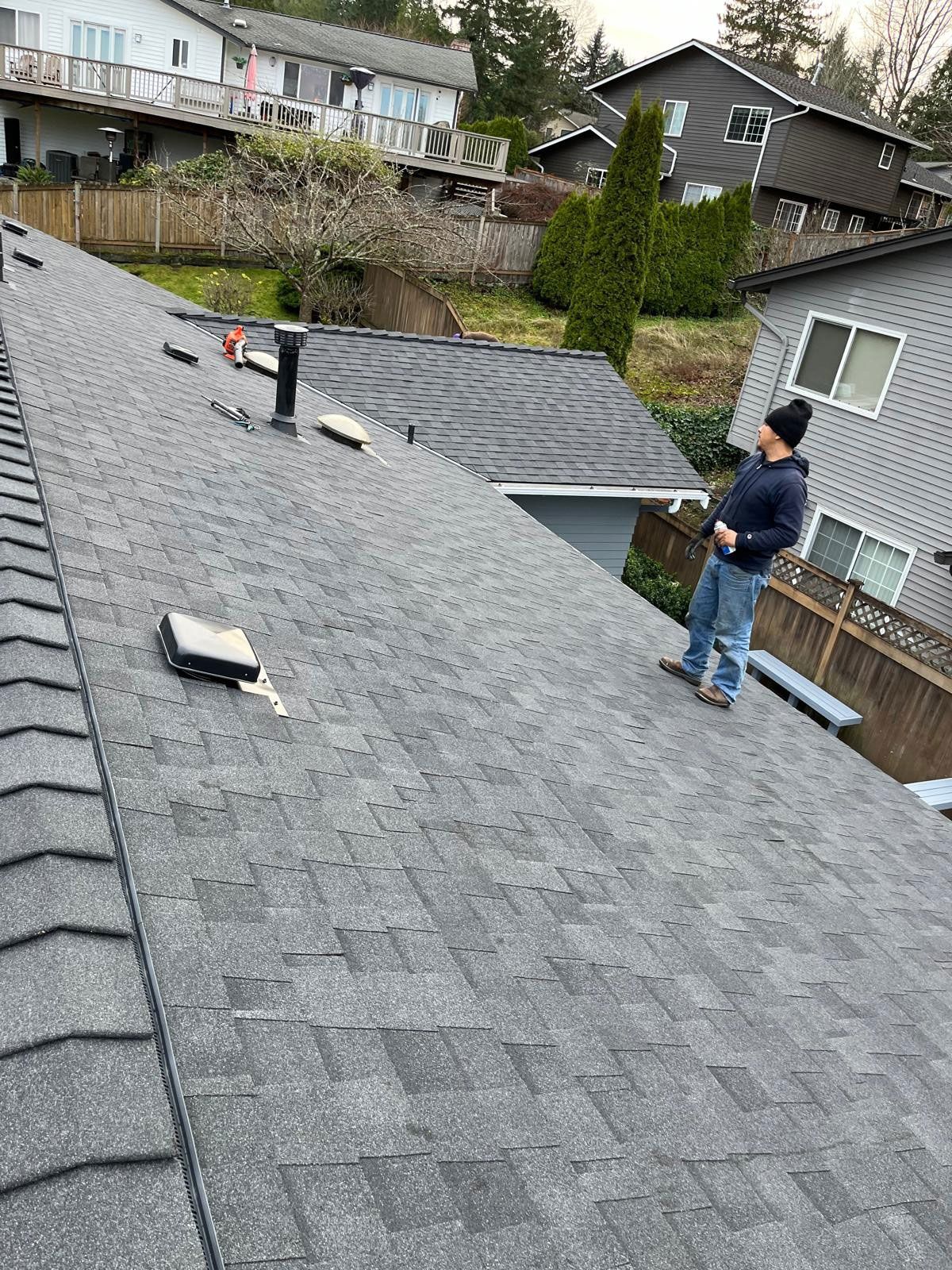 Person on a gray shingled roof, looking at another roof. Buildings and trees in background.