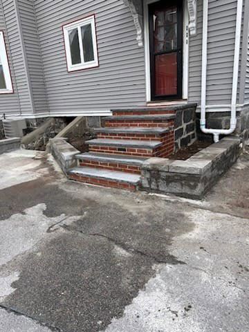 Brick steps leading to a gray house's red door, with a cracked concrete walkway.