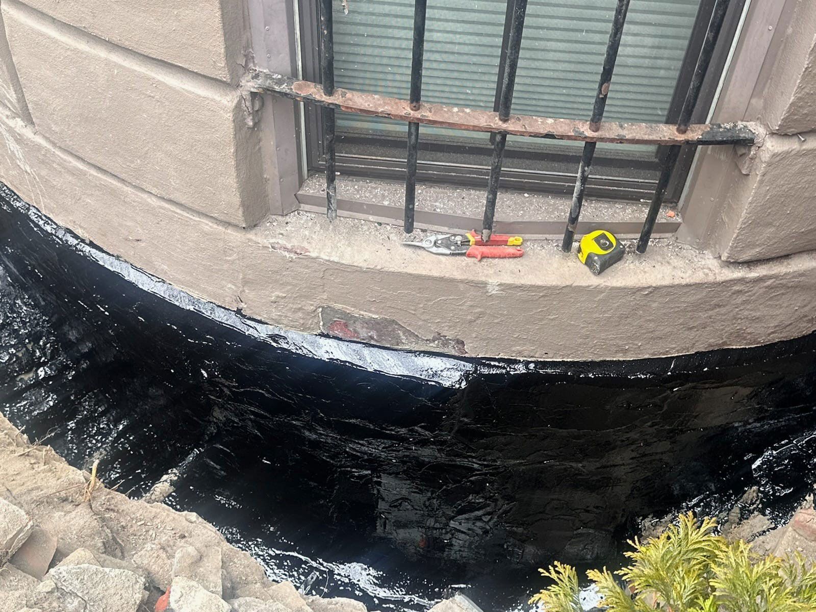 Black sealant applied around a window base, on a concrete structure. Tools visible nearby.