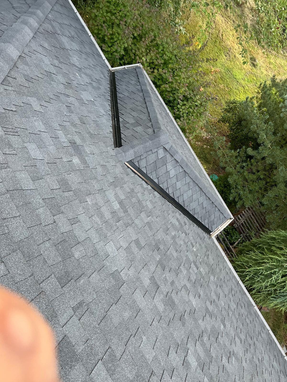Gray shingled roof with a black metal trim, viewed from above, with greenery in the background.