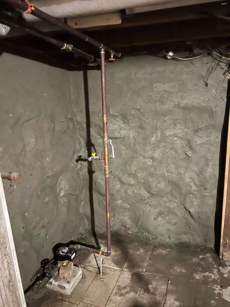 A basement with a concrete wall and copper pipes. Light shines on the wall.