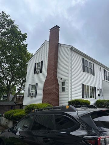 Two-story white house with a tall brick chimney, black shutters, and a car in front. Overcast sky.