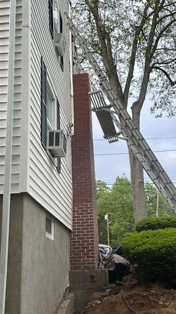 Brick chimney next to a white house. A worker is at the base; a tall ladder leans against it.