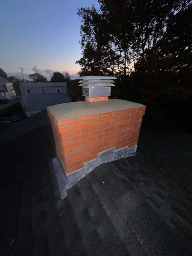 Brick chimney on a dark roof with a metal cap. Trees and buildings are visible in the background against a twilight sky.