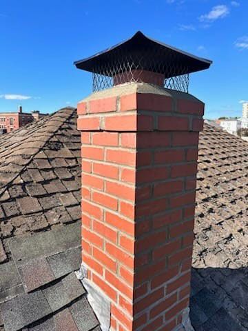 Brick chimney with a metal cap on a rooftop, against a blue sky.
