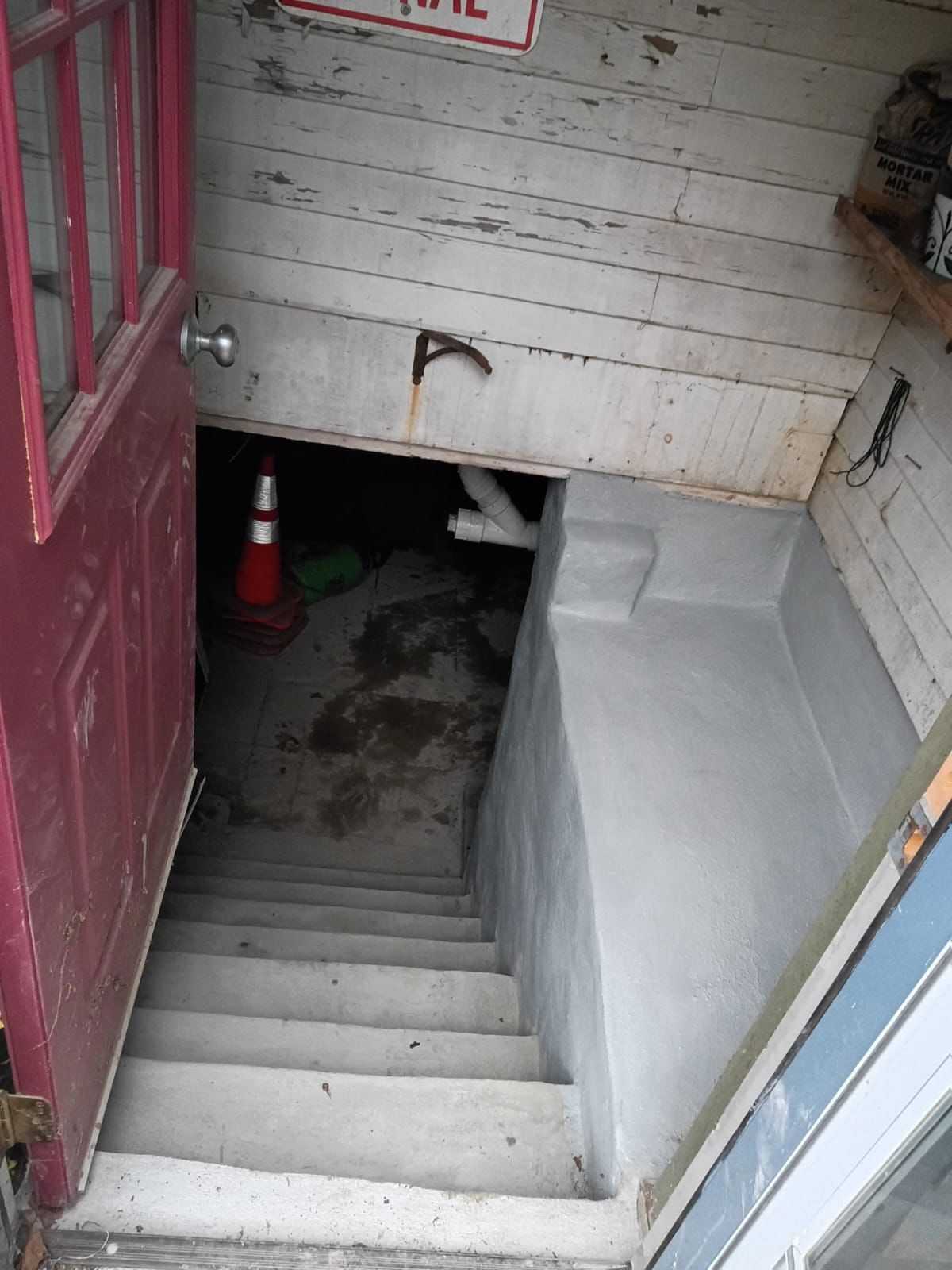 Staircase leading down from a red door. Concrete stairs and gray walls. A traffic cone is in the dark opening.