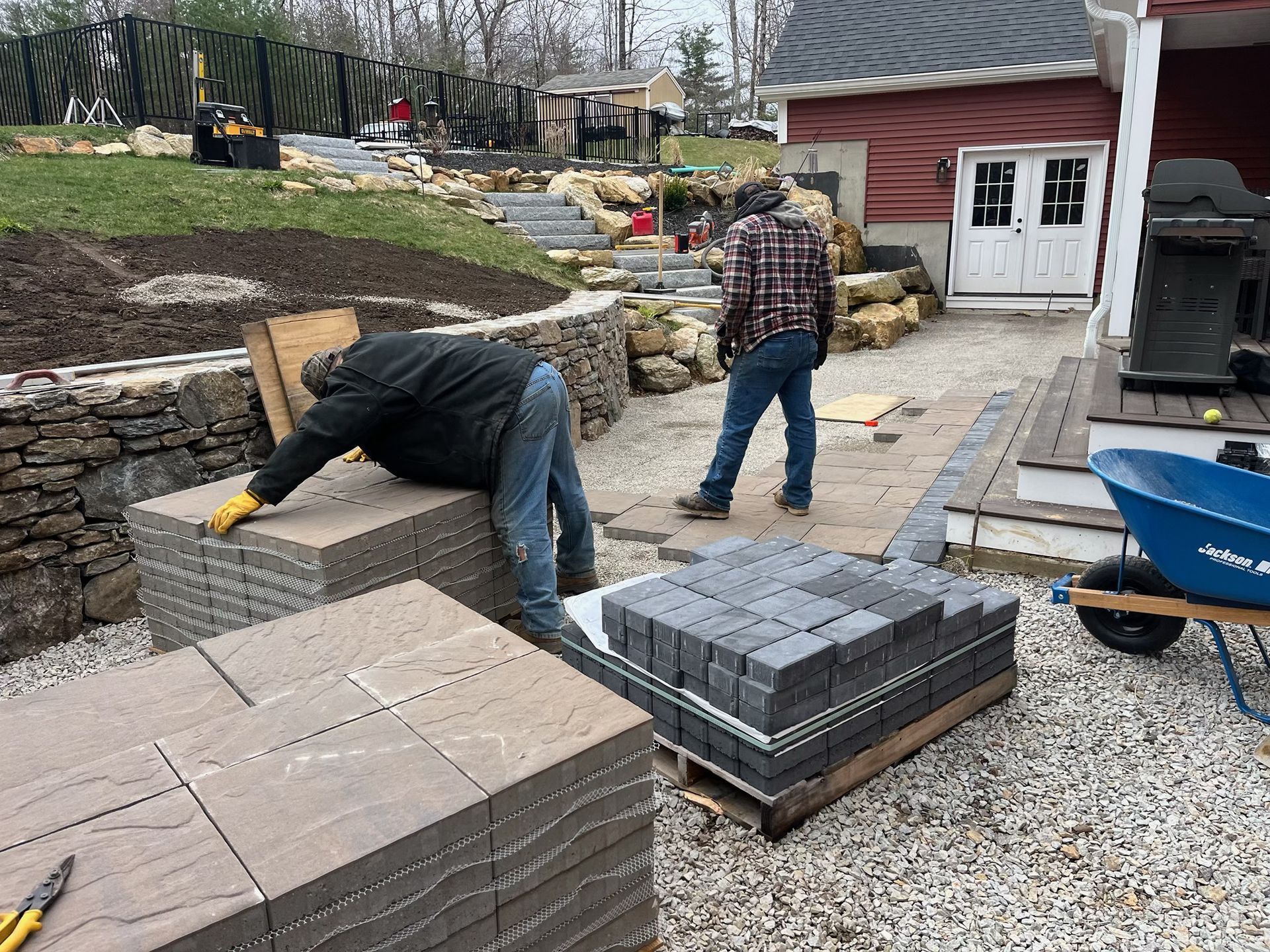 Two men are working on a patio with bricks and a wheelbarrow.