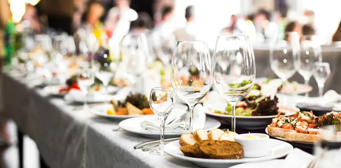 Long banquet table set with food and wine glasses, blurred figures in background.