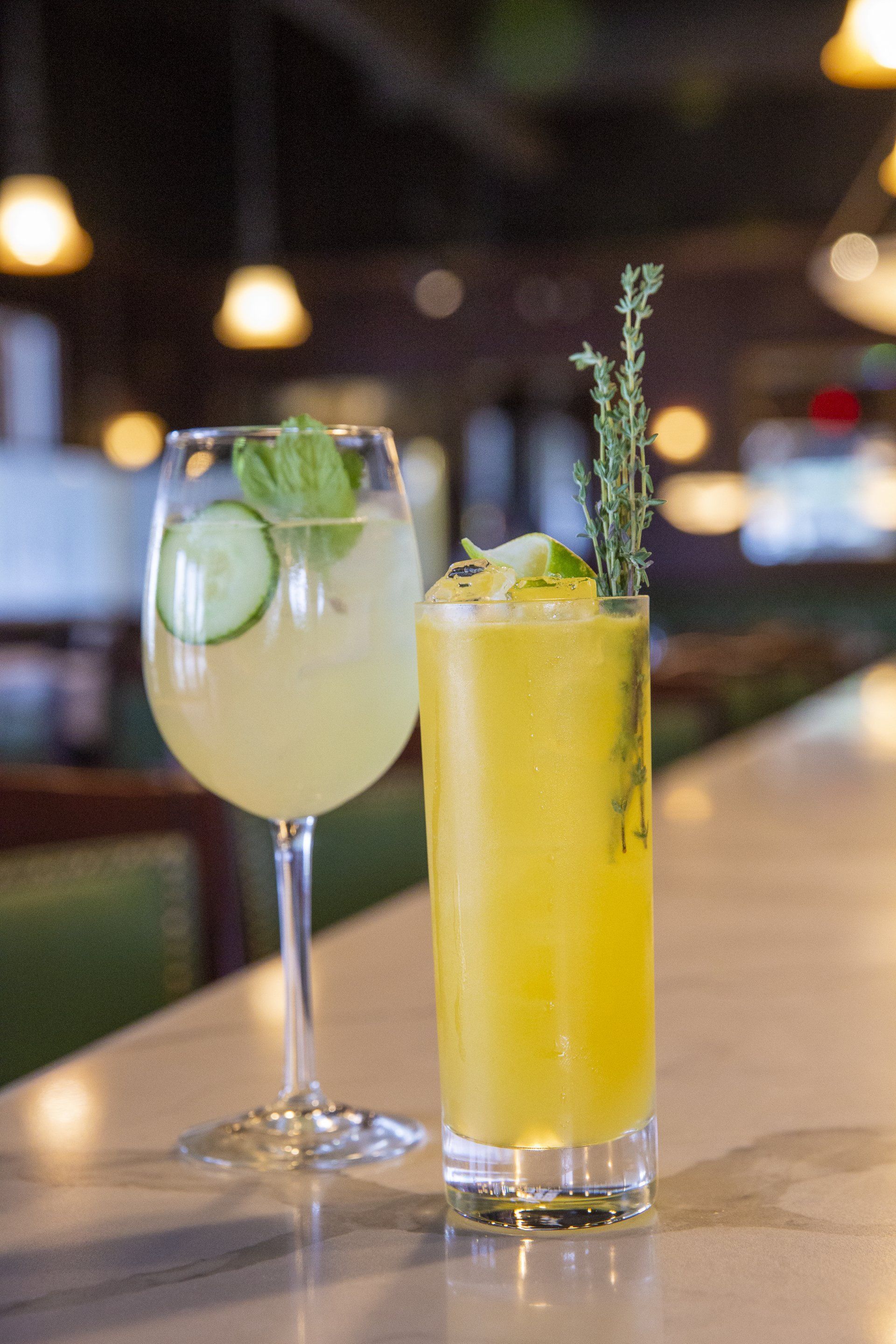 Two cocktails: one yellow, garnished with thyme, the other clear with cucumber in a bar setting.