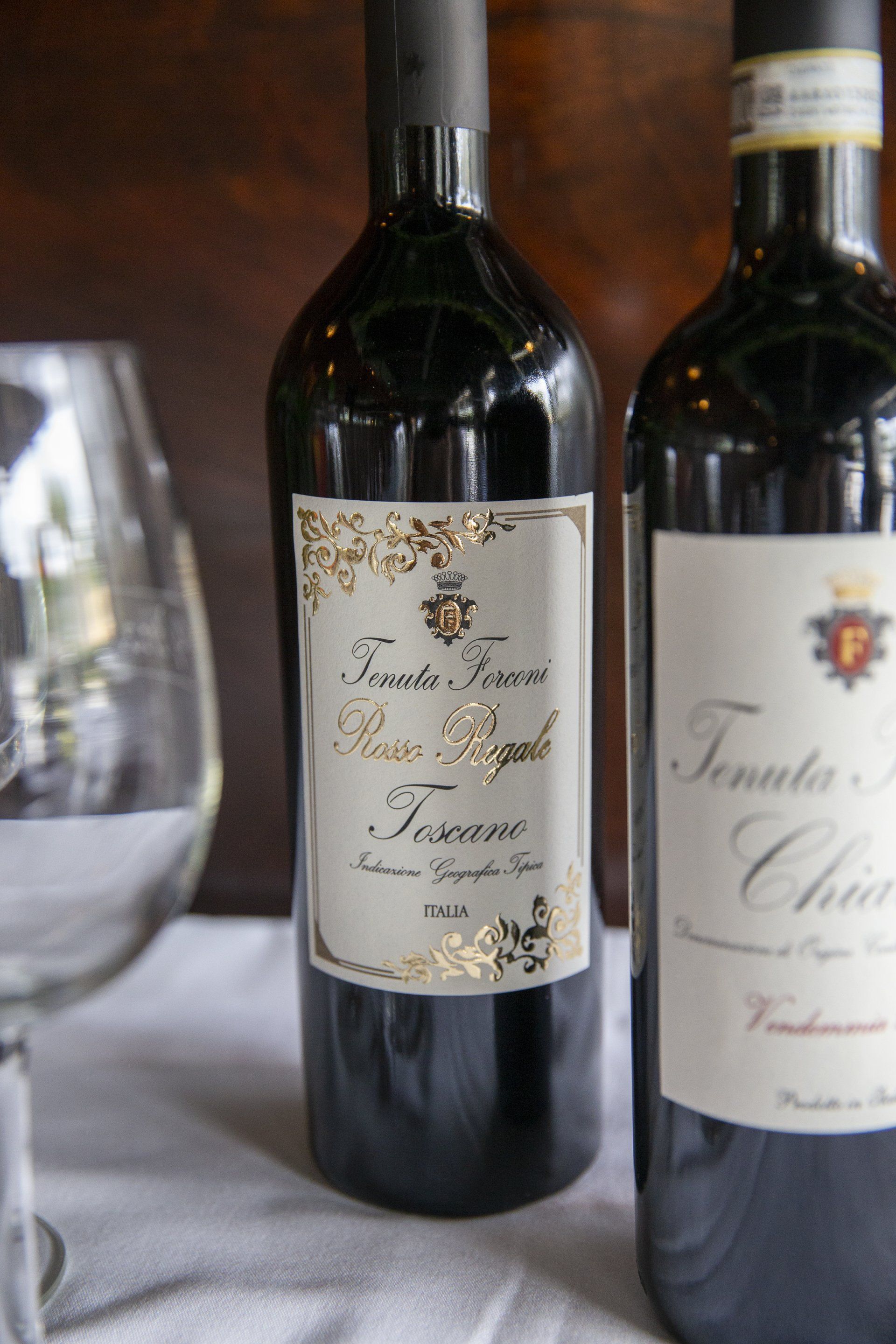 Two dark wine bottles on a table with a wine glass. 