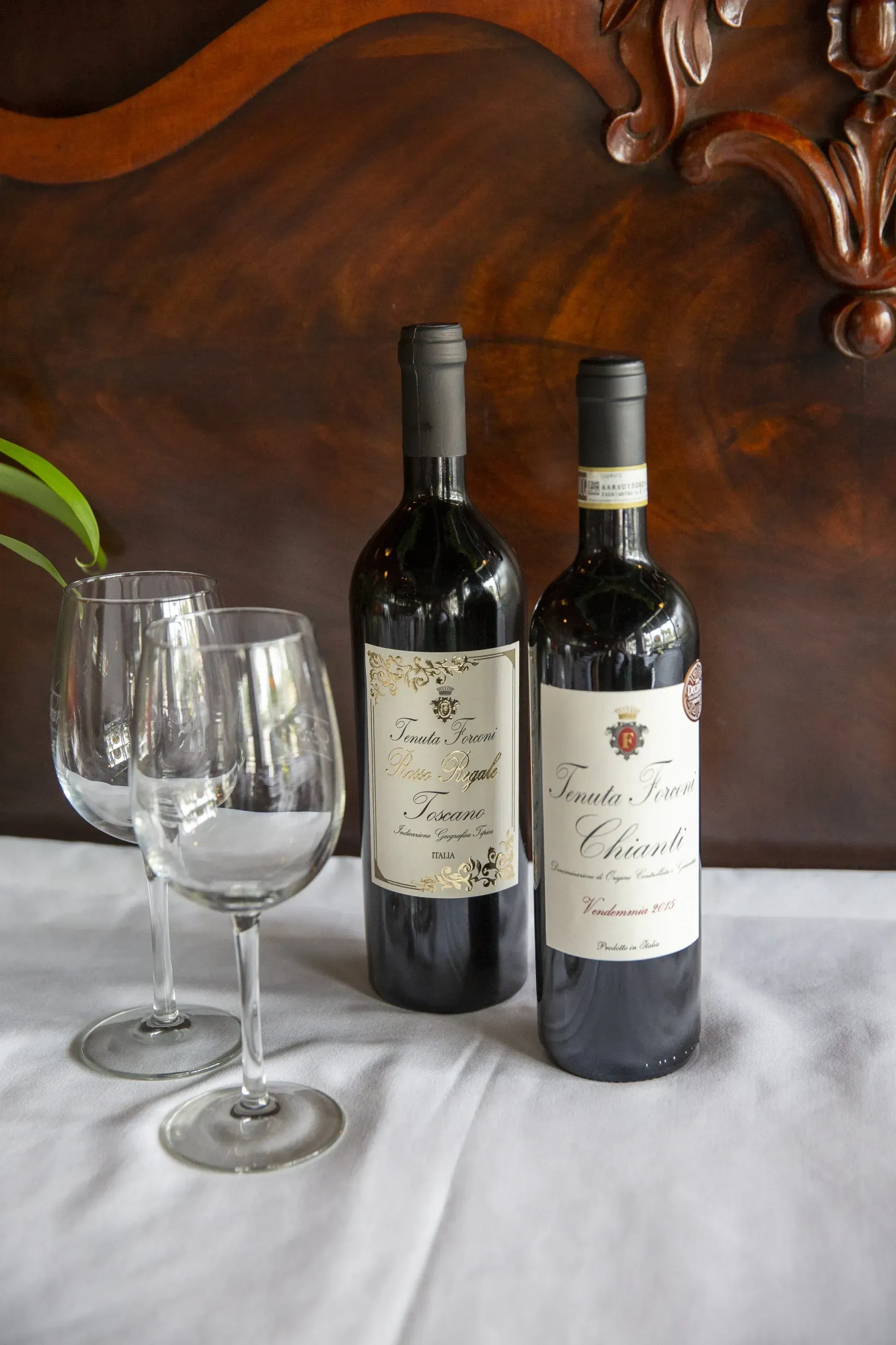 Two red wine bottles and two empty wine glasses on a white tablecloth, against a dark wood background.