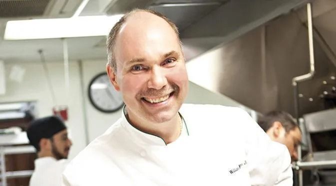 Chef smiles in kitchen, wearing white chef's coat; other staff visible in background.