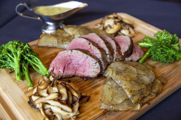 Sliced beef tenderloin with vegetables, mushroom, and sauce on a wooden platter.
