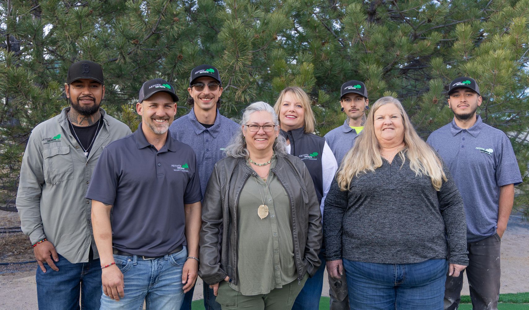 A group of eight people wearing casual clothing and hats stands together outdoors against a background of evergreen trees.