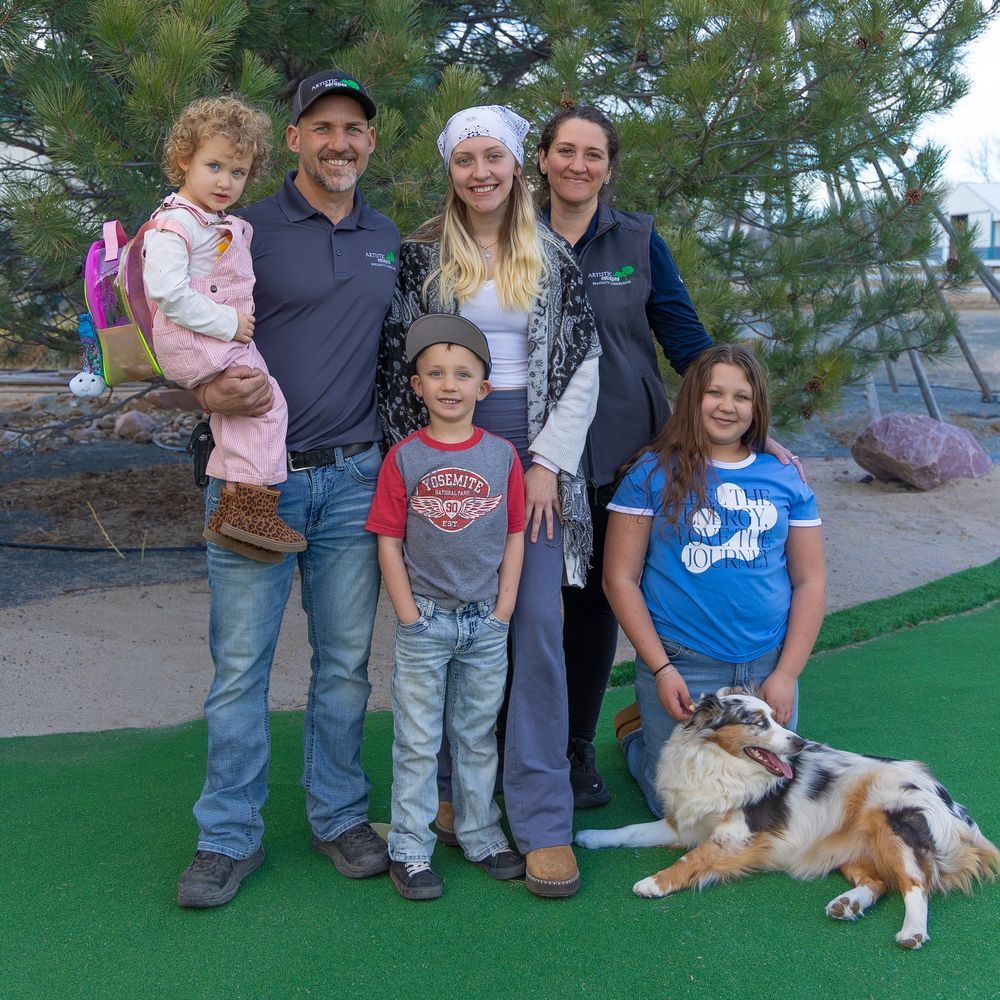 A family of six and their dog stand together on a green turf area outdoors, posing for a portrait in front of pine trees.