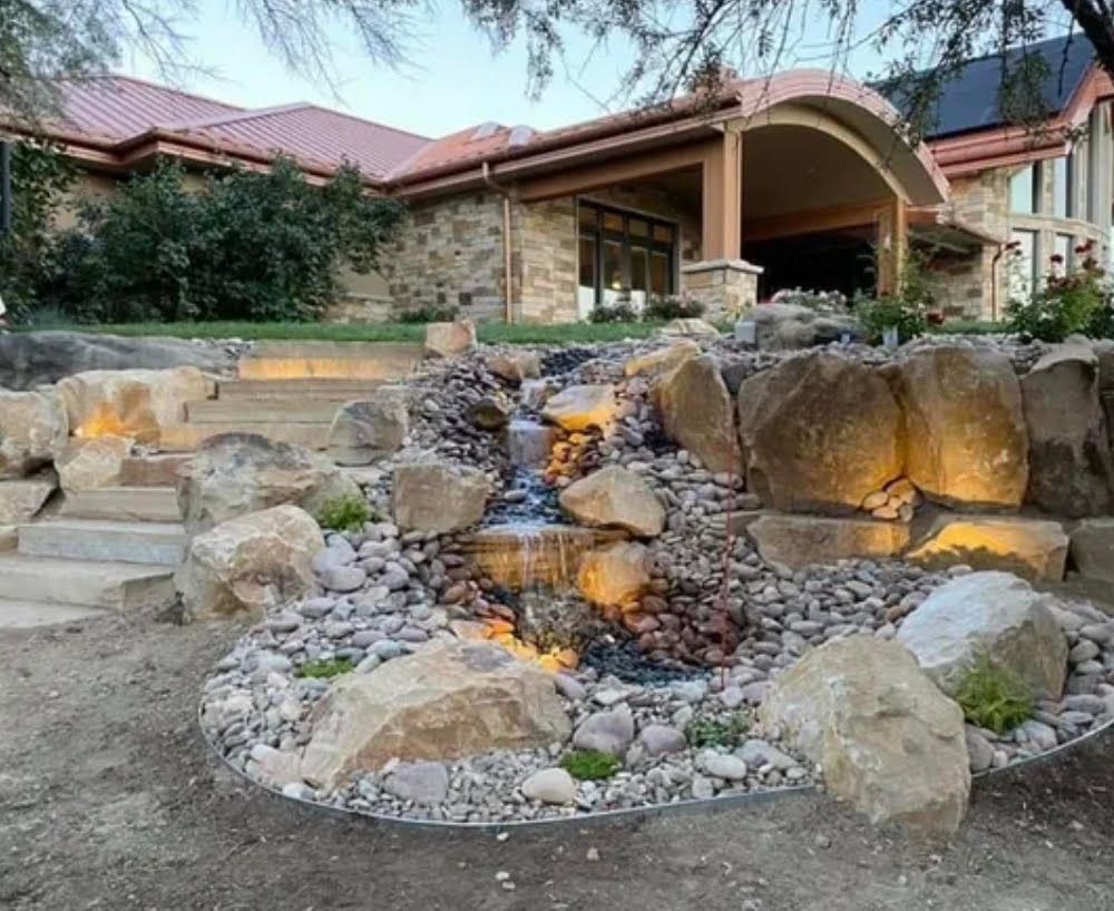 Rock garden waterfall with lit stones in front of a home with a curved porch