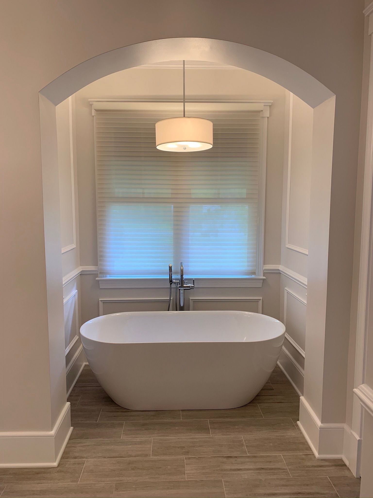 Freestanding white bathtub in an alcove with a pendant light and window, neutral color palette.