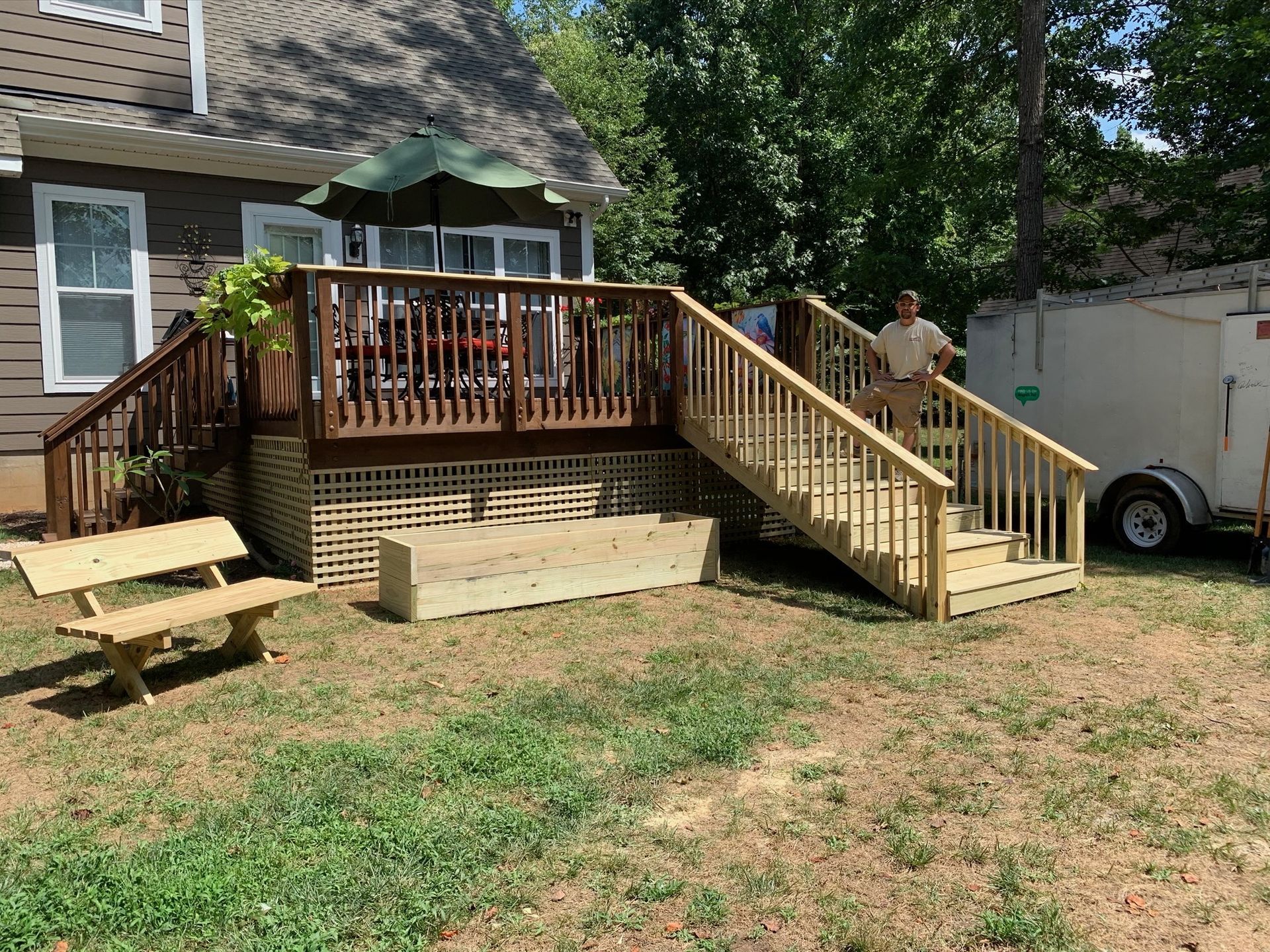 Backyard deck with stairs; man stands on steps. Brown house, bench, umbrella.