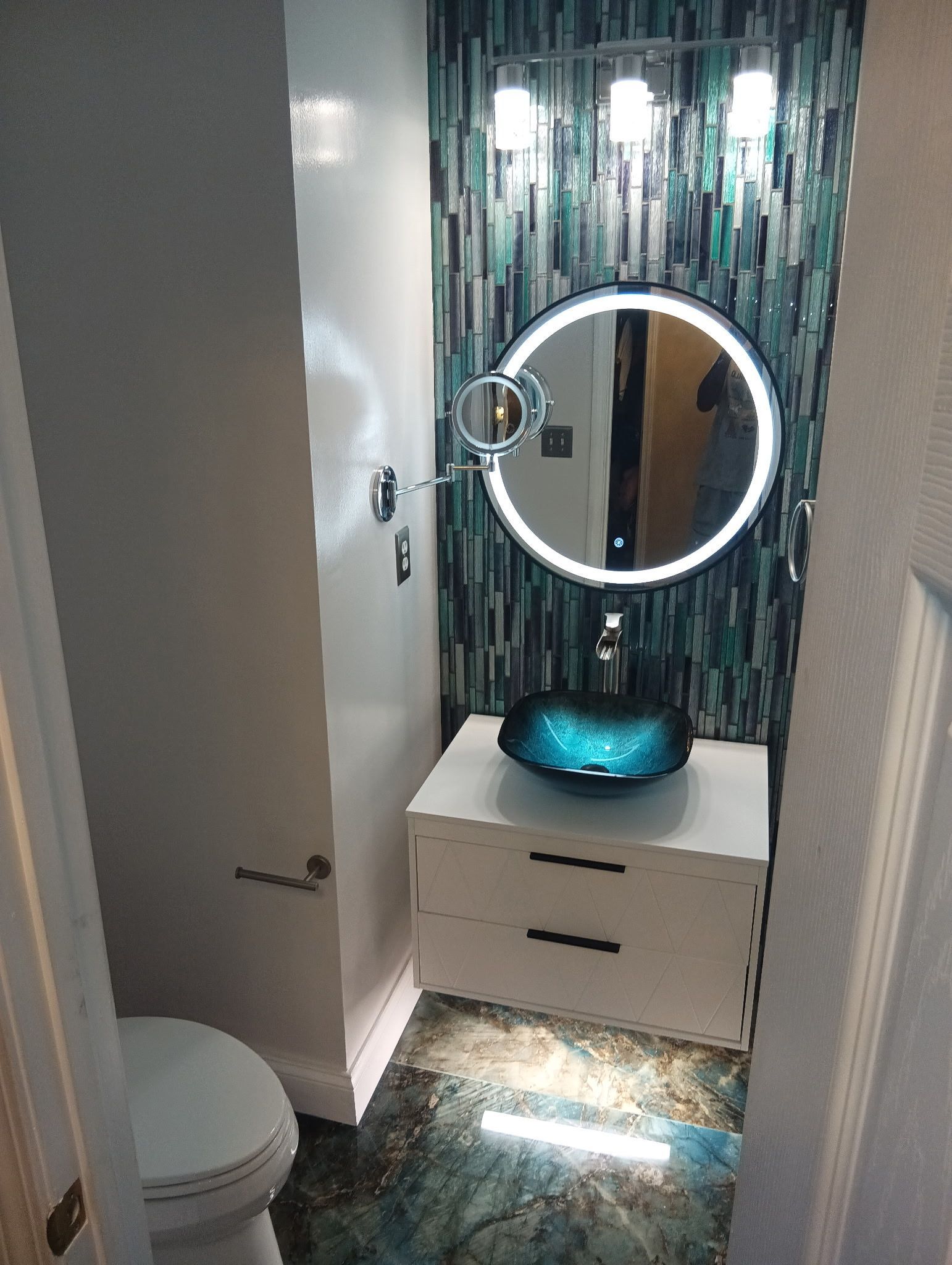 Small bathroom with mosaic tile accent wall, blue glass vessel sink, round mirror, and white cabinet.