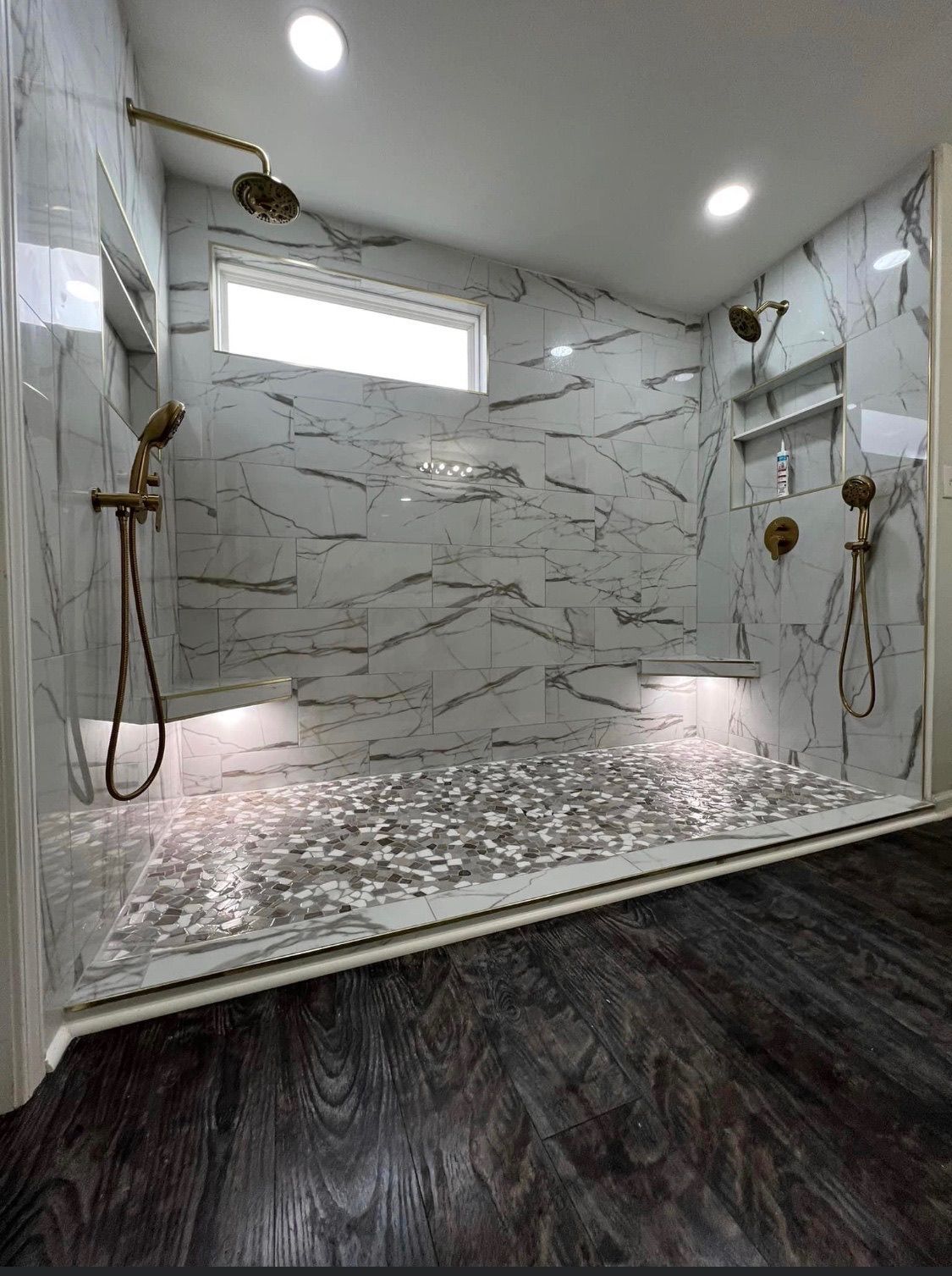 Luxurious walk-in shower with marble-like tiles, multiple showerheads, and a mosaic floor. Dark wood flooring.