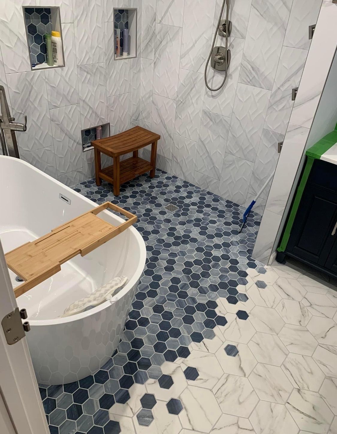 Bathroom with a freestanding tub, wooden bench, and blue-gray hexagonal floor tile. White marble-look wall tile.