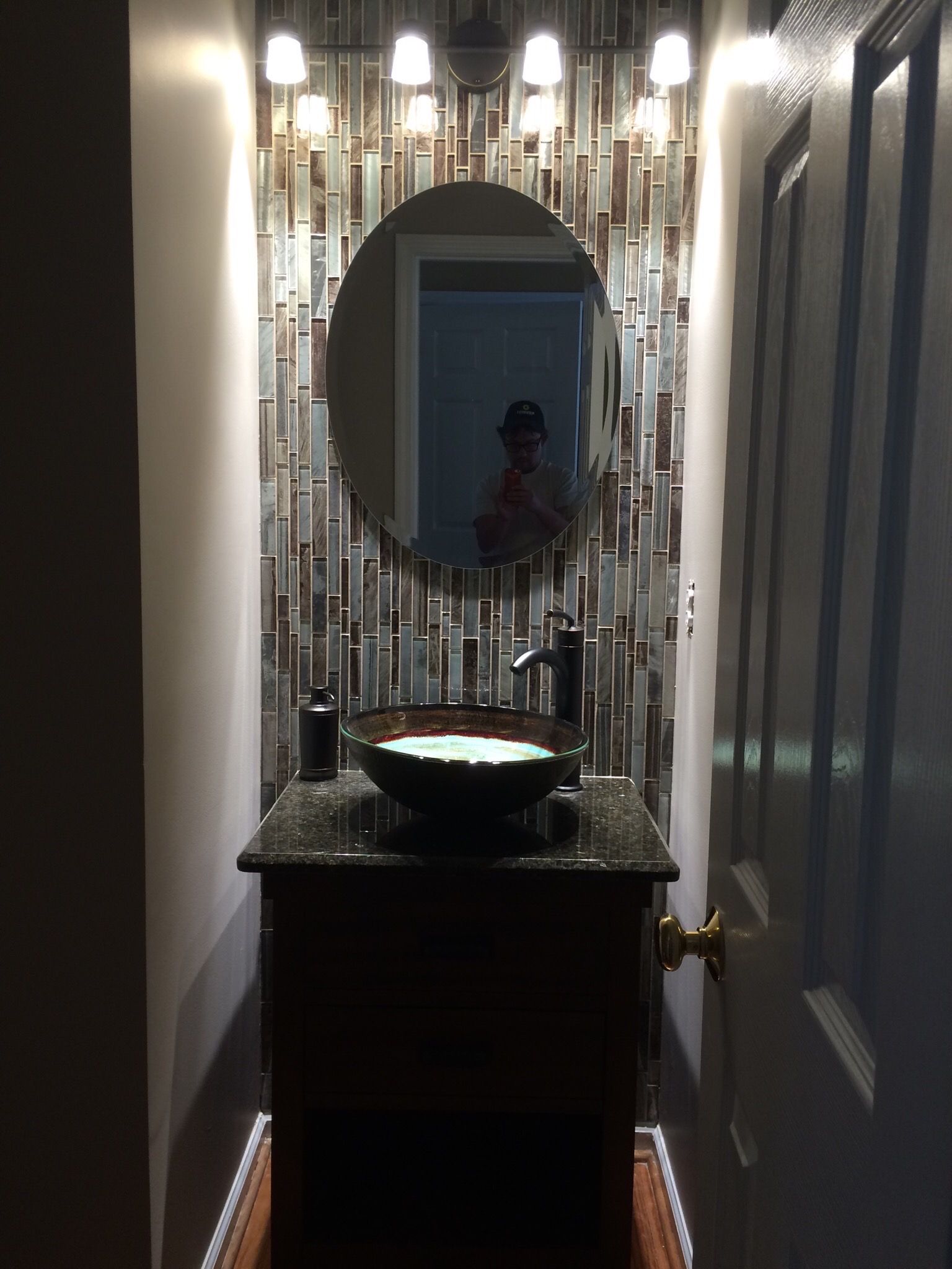 Small bathroom with dark cabinet, mosaic tile backsplash, round mirror, and vessel sink.