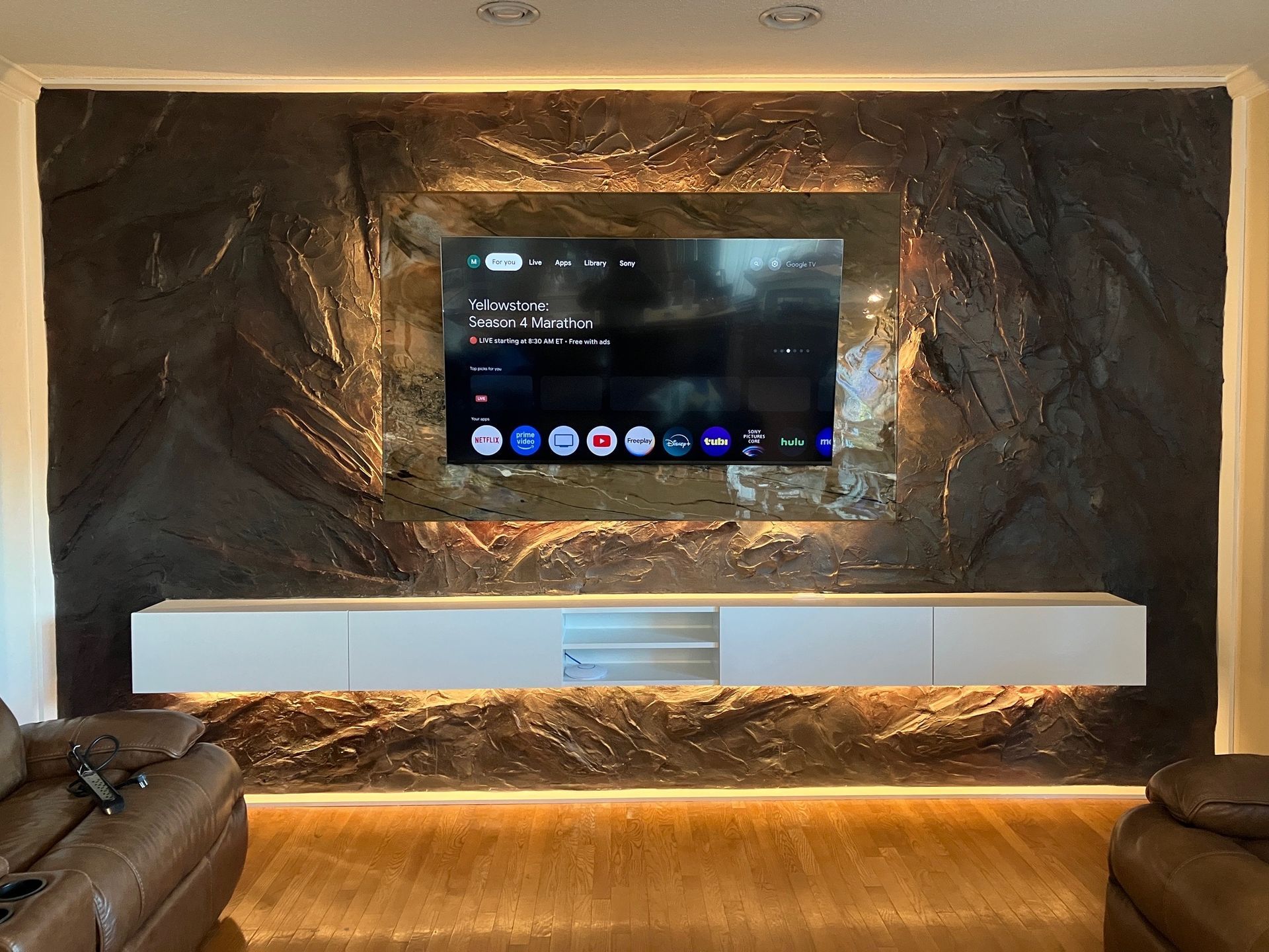 A modern media wall with a large TV centered on a dark stone feature wall, floating white cabinet, and ambient lighting.