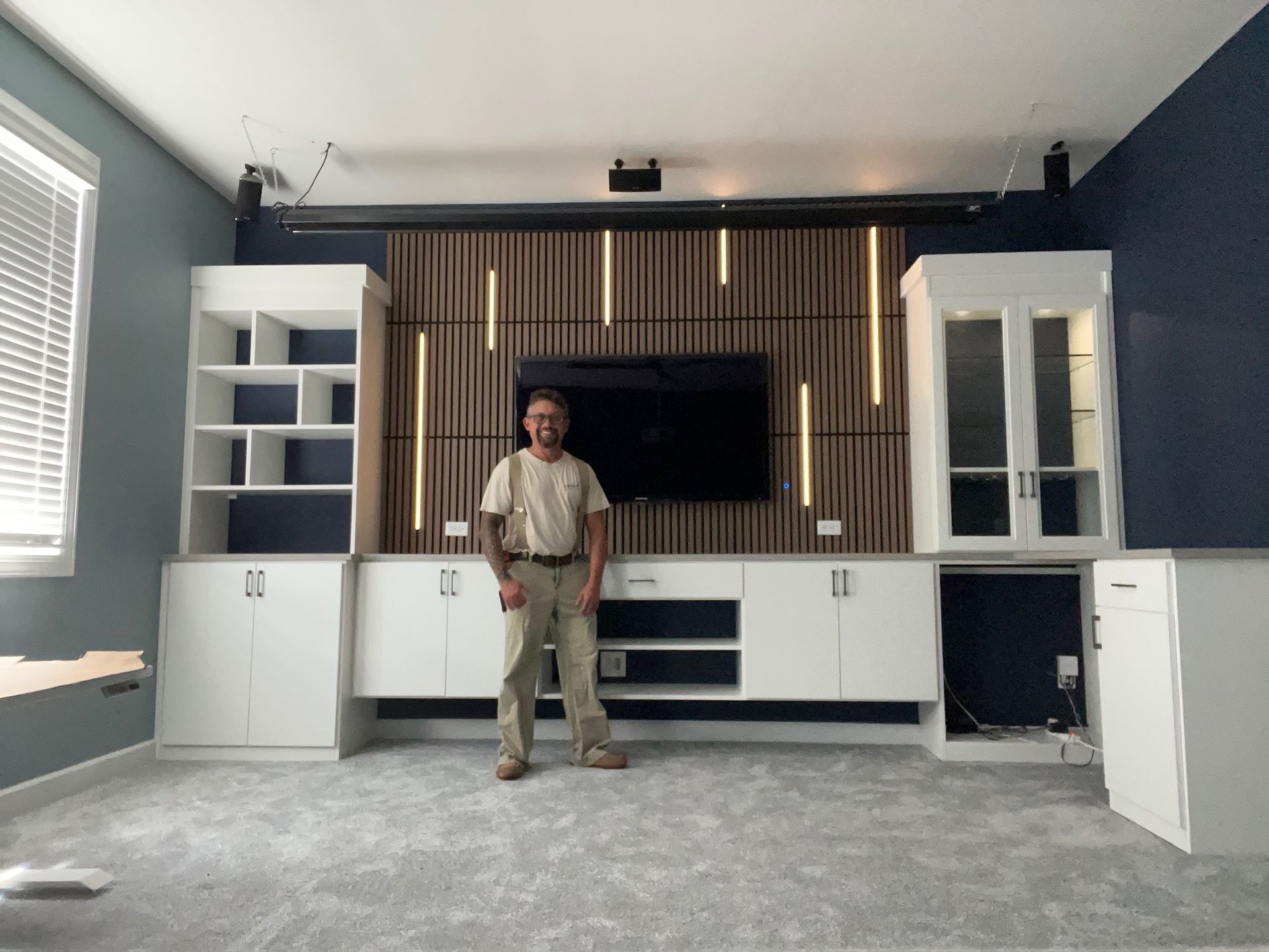 Man stands in front of a custom entertainment center with a TV, shelves, and lights. The walls are gray and blue.