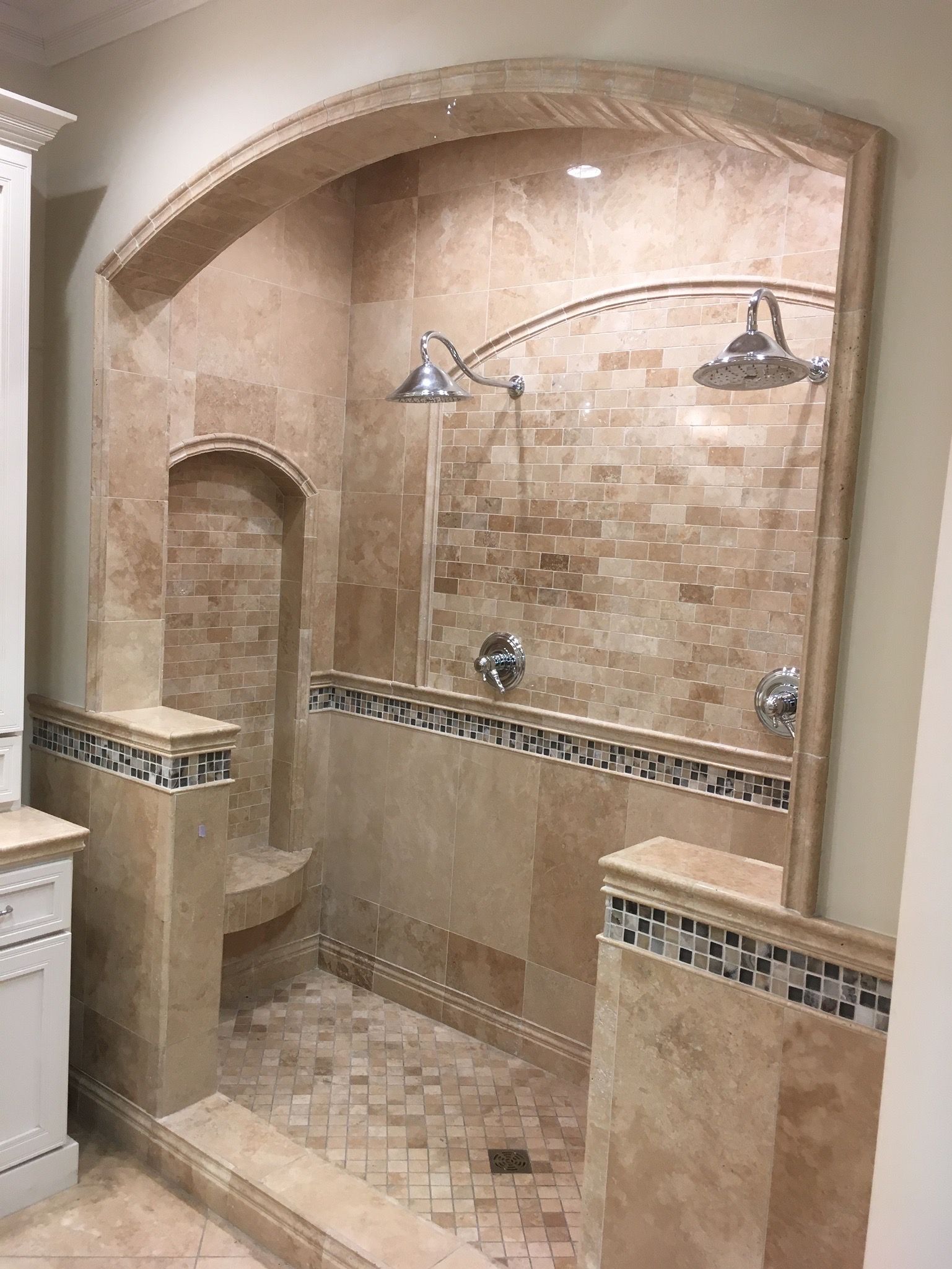 Spacious, beige tile shower with built-in seating and dual showerheads. Archway and mosaic tile accents.